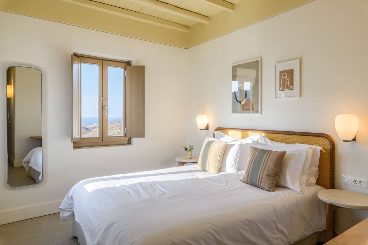 A bedroom with a bed, a bedside table, a mirror, a window with a view, and wall-mounted lights.