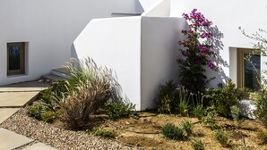 Un edificio moderno blanco con un diseño geométrico, un pequeño jardín con piedras y plantas, y un cielo azul despejado.