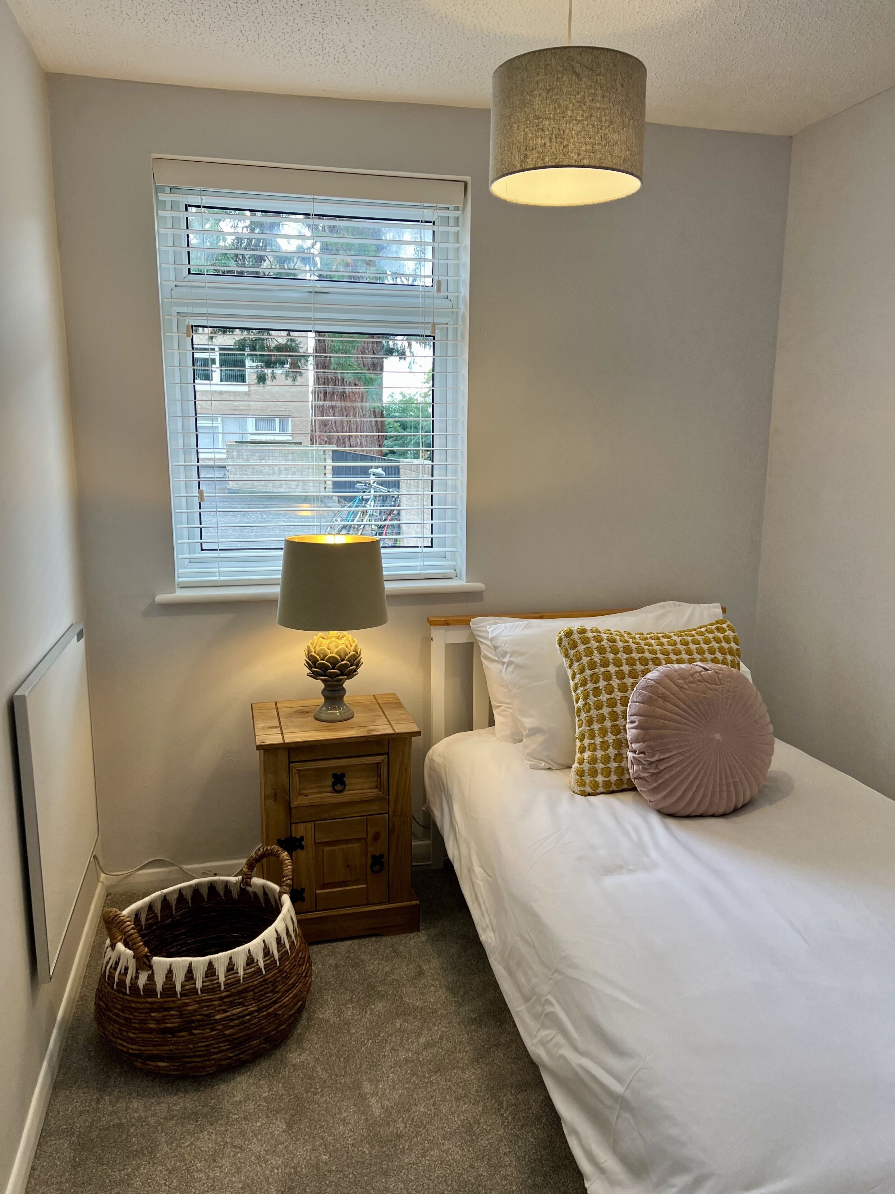 Un dormitorio con cama, mesita de noche con lámpara, ventana con persianas y una cesta de mimbre.