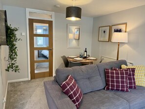 A living room with a grey sofa, a dining table set with plates and glasses, a lamp, and a door leading to a bedroom. - BookedUK - Lovely 2BR Flat in Hoddesdon (Hoddesdon)