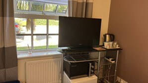 A room with a television on a stand, a small refrigerator, a radiator, and a window with curtains. - SleepNeat (Ascot)