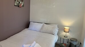 A neatly made bed with white linens, a small bedside table with a lamp, and a trash can. - SleepNeat (Ascot)