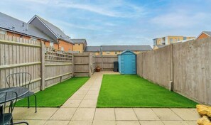 Un jardin à l’arrière d’une maison, avec une terrasse dallée, de l’herbe artificielle verte, une clôture en bois et un abri de jardin bleu.