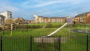 A playground with a slide and climbing structures, surrounded by a green lawn and a black fence. - 4BR-Madejski Stadium-Royal Berk Hosp-By Tempstay (Reading)