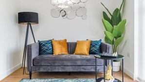 A grey sofa with blue and yellow patterned cushions, a black floor lamp, a large green plant, and a chessboard on a small table. - 4BR-Madejski Stadium-Royal Berk Hosp-By Tempstay (Reading)