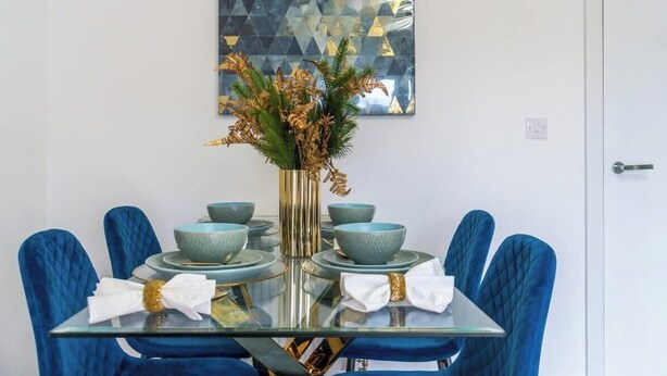 A dining table with blue chairs, a glass top, and a vase with dried flowers. - 4BR-Madejski Stadium-Royal Berk Hosp-By Tempstay (Reading)