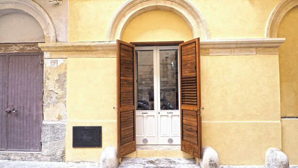 A yellow building with a wooden door and shutters, a stone plaque, and a stone base. - Corte Tiso (Lecce)