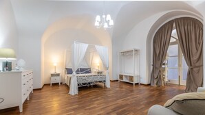 A bedroom with a four-poster bed, a nightstand, a dresser, a bookshelf, and a large arched window with curtains. - Corte Tiso (Lecce)