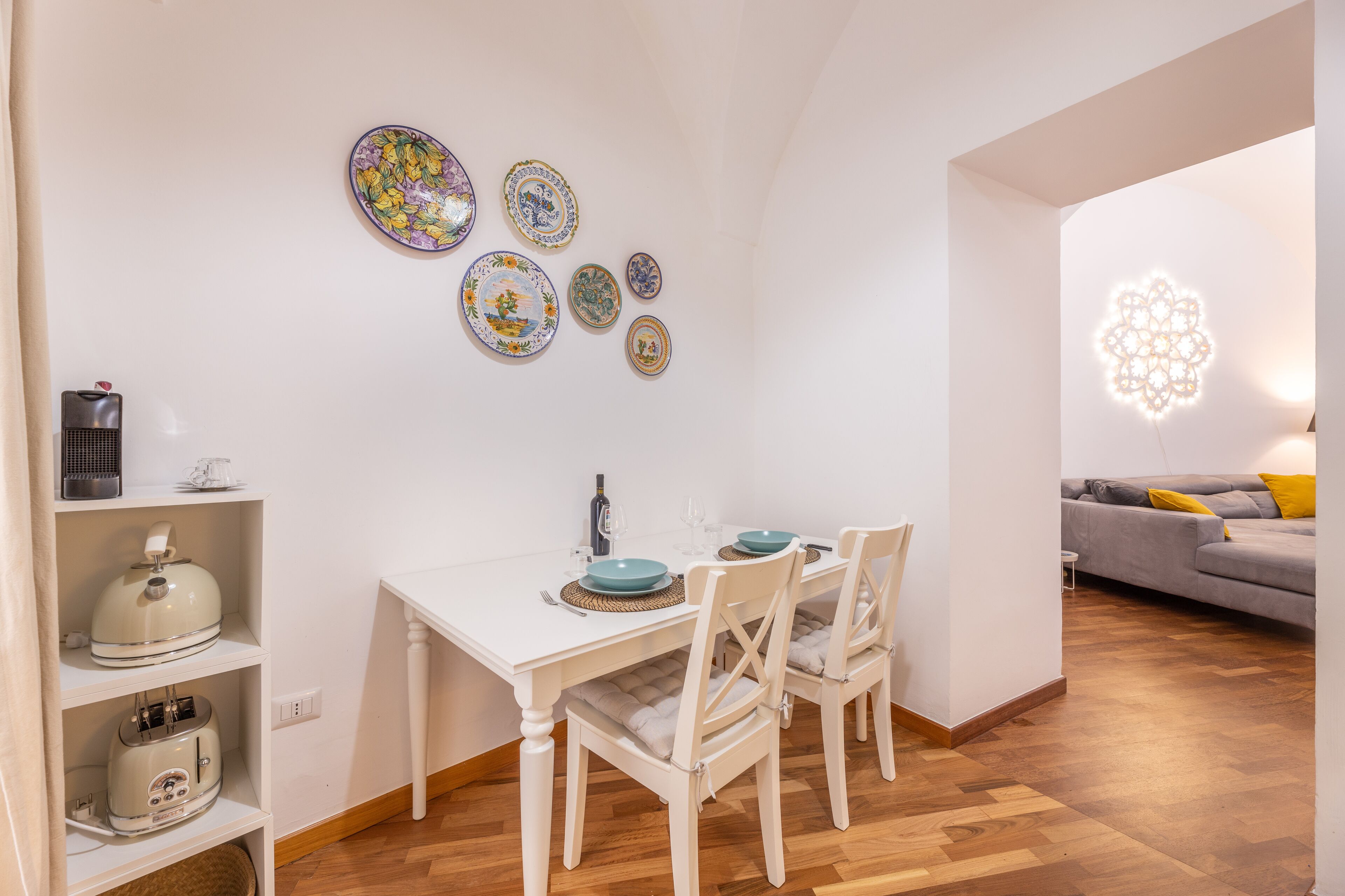 Un'area pranzo con un tavolo bianco apparecchiato per due, circondato da sedie bianche. Sulla parete sopra il tavolo sono presenti piatti decorativi.
