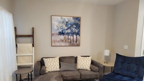 A living room with a sofa, ottoman, and a blue chair. There is a painting on the wall and a lamp on a side table. - New Branson Condo!! (Branson)