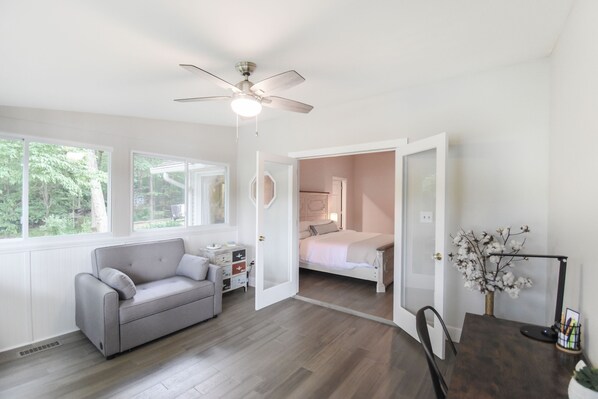 A room with a grey sofa, a wooden dining table, a ceiling fan, and a bed with a white headboard.