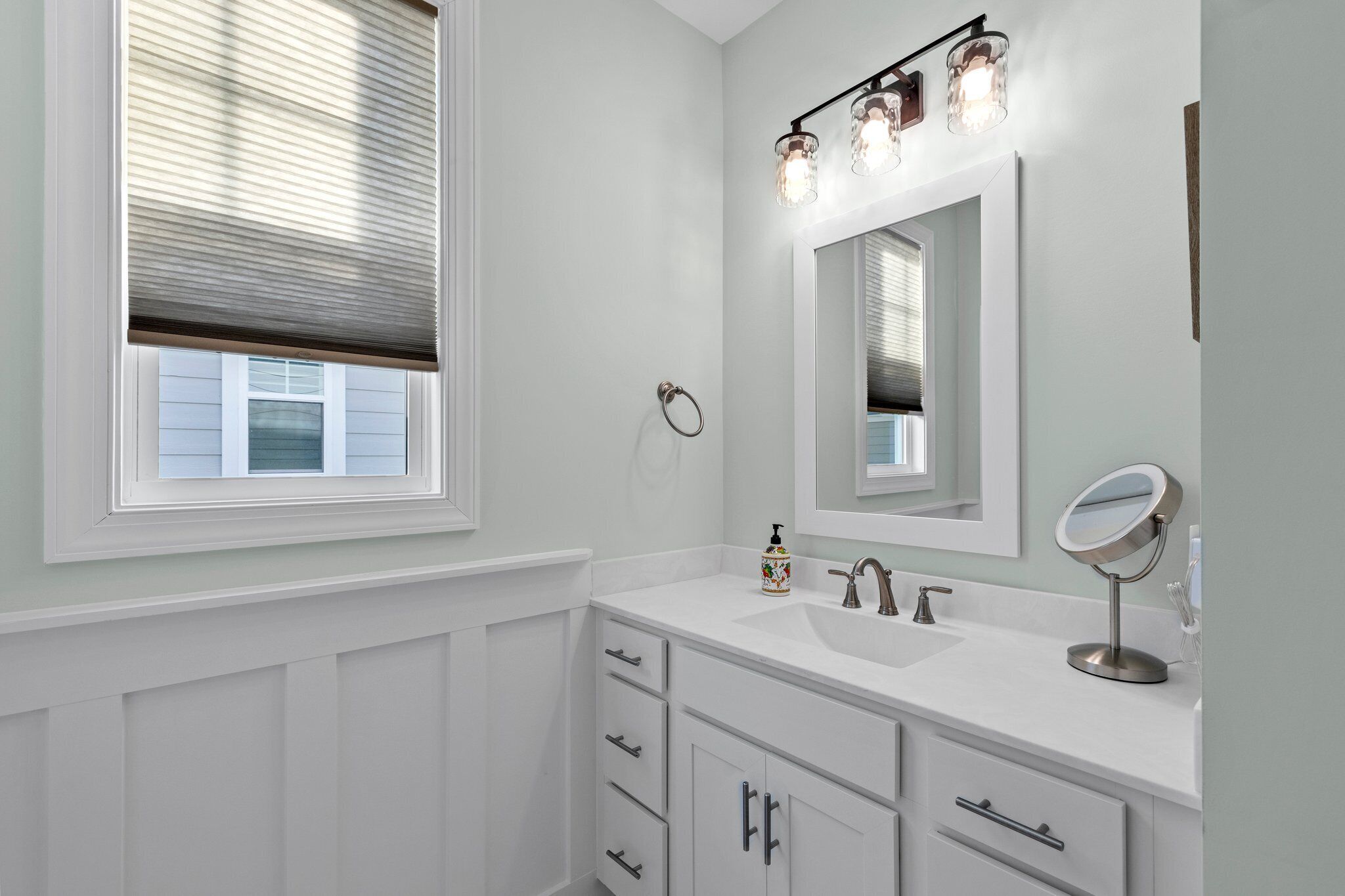 A bathroom with a window, white vanity, mirror, and light fixture.