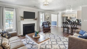 A spacious living room with a fireplace, a TV mounted above it, and a dining area with bar stools. - Spacious King Suite w/ Game Room, Fire Pit & Patio (Lexington)