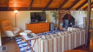 Ein gemütliches Wohnzimmer mit Holzbalkendecke, einem Fernseher, einer Couch, einem Sessel und einem Kamin.