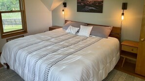 A bedroom with a large bed, two bedside tables, a nightstand, a window with a view of greenery, and a sunset painting on the wall.