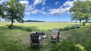 An outdoor seating area with two chairs and a fire pit, overlooking a grassy field and a body of water.