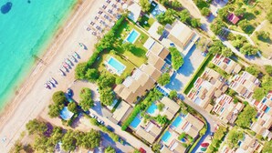 Vista aérea de un complejo turístico costero con alojamiento frente a la playa, piscina y exuberante vegetación.