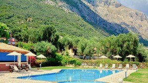 Zona de piscina con sillas de descanso y sombrillas, rodeada de exuberante vegetación y montañas.