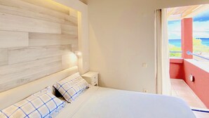 Un dormitorio con cabecera de madera, una mesita de noche con lámpara y vista al mar a través de una puerta abierta.