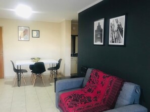 A living room with a blue sofa and a dining table with four chairs. There are two framed pictures on the wall.