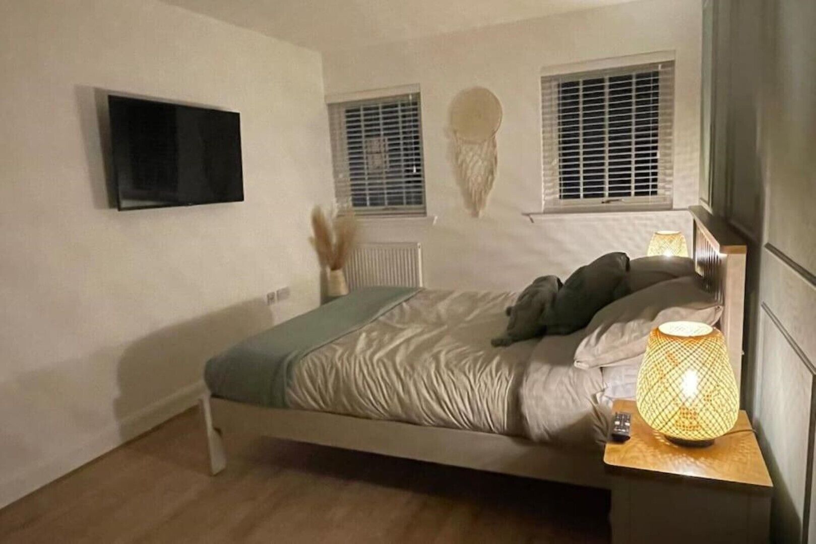 A bedroom with a bed, a bedside lamp, a TV mounted on the wall, and two windows with blinds.