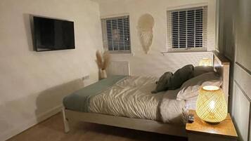A bedroom with a bed, a bedside lamp, a TV mounted on the wall, and two windows with blinds.