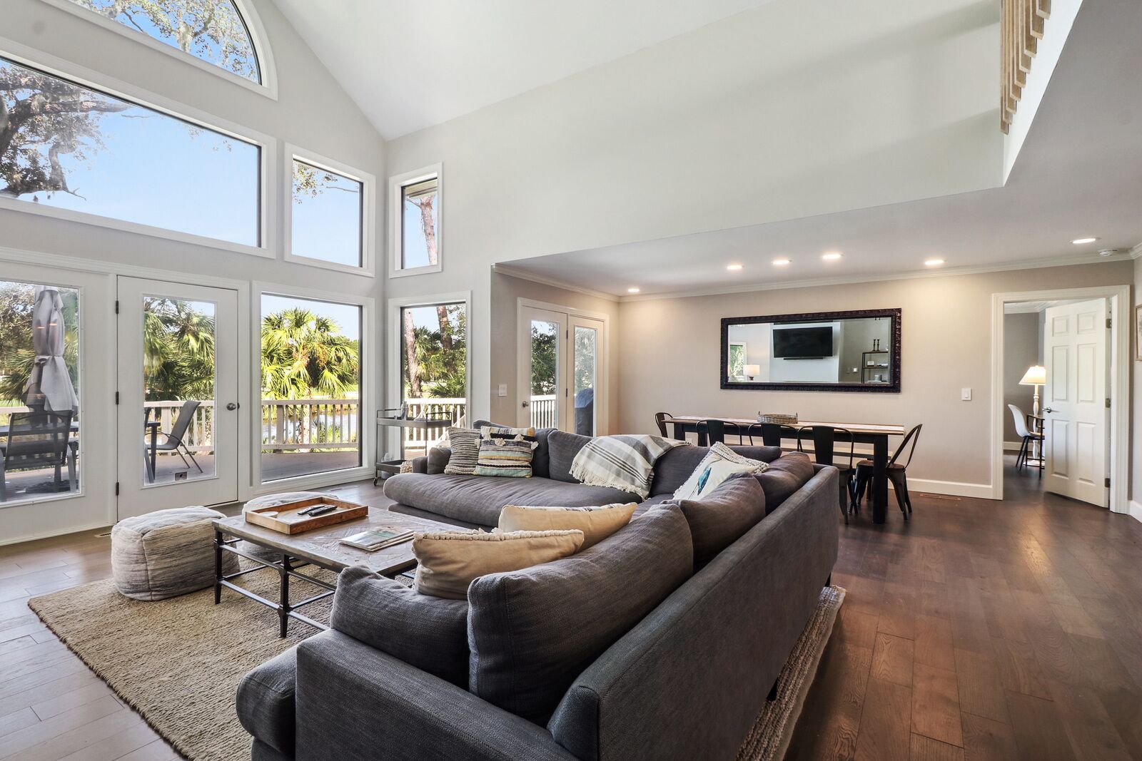 A spacious living room with a large gray sectional sofa, a dining area with a table and chairs, and a large TV mounted on the wall.