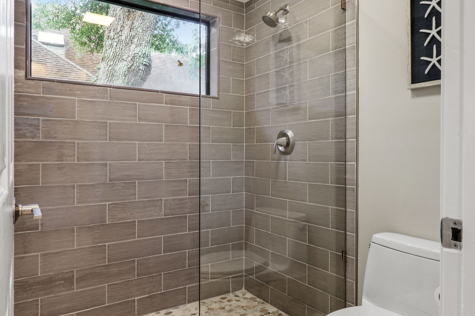A modern bathroom with a glass shower enclosure, a toilet, and a window with a view of trees and a building.