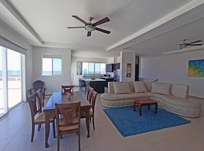 Ampla sala de estar com mesa de jantar, cadeiras, sofá e tapete azul. Há ventiladores de teto, uma cozinha ao fundo e janelas grandes com vista.