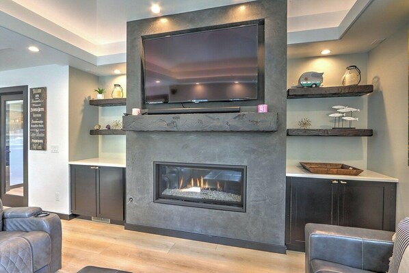 A modern living room with a flat-screen TV mounted above a fireplace, wooden shelves with decorative items, and leather seating.