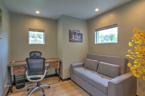 A room with a desk, office chair, and a sofa. There are two windows and a painting on the wall.