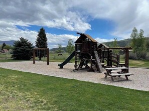 Une aire de jeux dotée d’une structure en bois, comprenant un toboggan et une zone d’escalade, entourée d’un chemin de gravier et d’espaces verts.