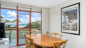 A dining area with a wooden table and chairs, large windows with a view of the beach, and a framed artwork on the wall.