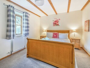 A bedroom with a wooden bed, two bedside tables with lamps, a radiator, a window with curtains, and a floral painting on the wall. - Waggoners Cottage-UK12901 (Pelynt, near Looe)