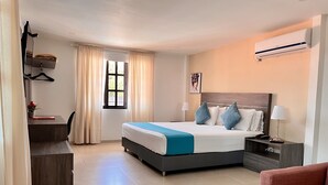 Habitación de hotel con cama, mesitas de noche, escritorio, silla, ventana con cortinas y equipo de aire acondicionado.