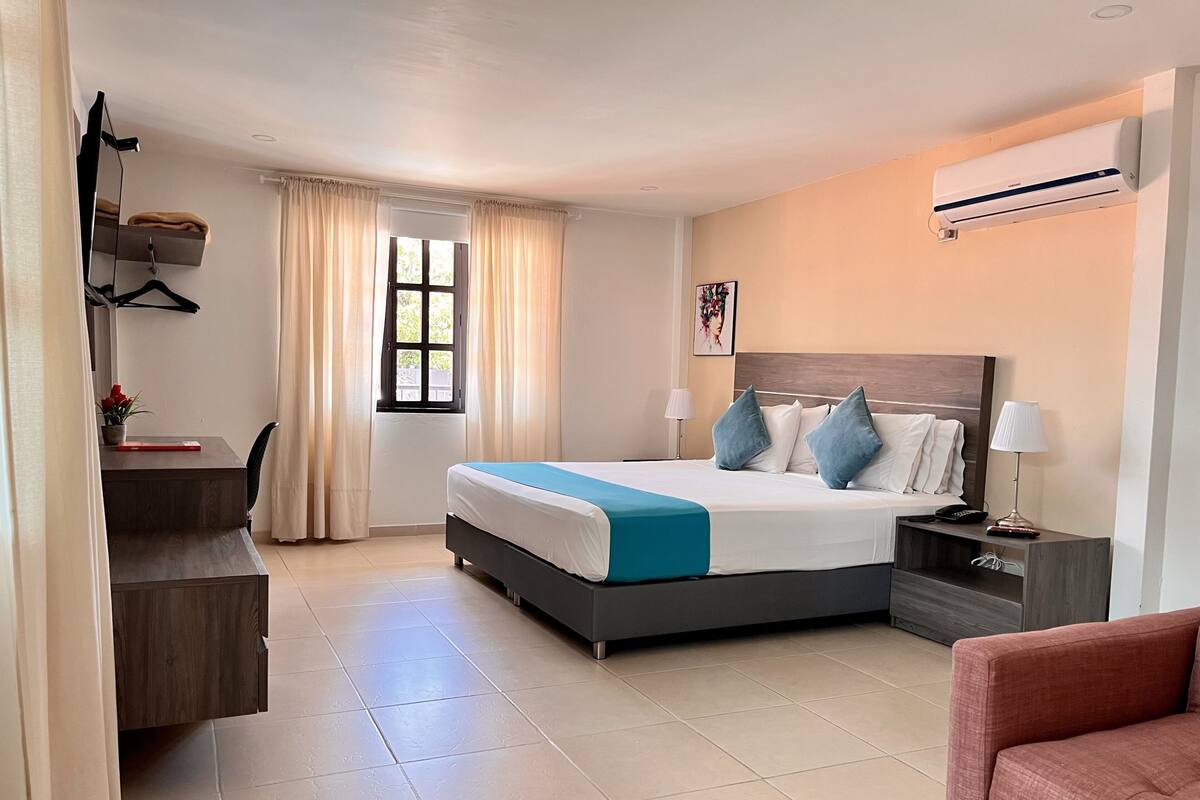 Une chambre d’hôtel comprenant un lit, des tables de chevet, un bureau, une chaise, une fenêtre avec des rideaux et un appareil de climatisation.