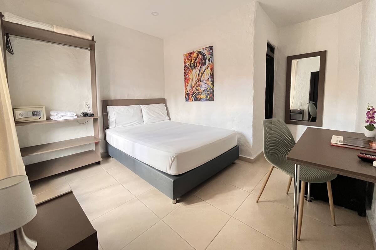 Une chambre d’hôtel comprenant un lit, un bureau, une chaise, une étagère avec un téléphone et une radio, un tableau au mur et un miroir.