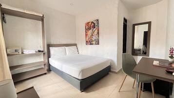 Quarto de hotel com uma cama, escrivaninha, cadeira, prateleira com telefone e rádio, um quadro na parede e um espelho.