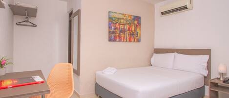 A hotel room with a bed, a desk, a chair, a painting, and an air conditioning unit.