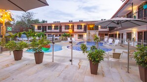 Un área de piscina con sillones de descanso y sombrillas, rodeada de plantas en macetas y un edificio con varios balcones.