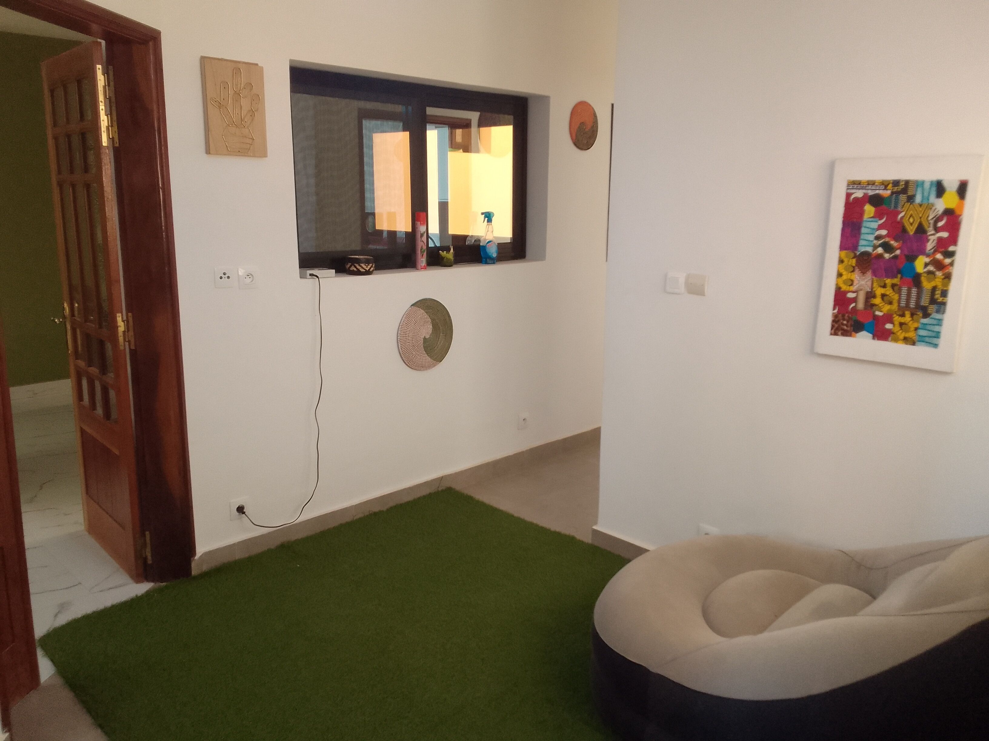 A room with a green carpet, a round wall decoration, a window with a mirror, and a colorful artwork on the wall.