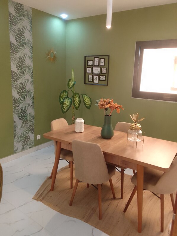 Un comedor con una mesa de madera, sillas color beige, un jarrón verde con flores y un cuadro en la pared.