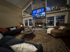 A living room with a stone fireplace, a flat-screen TV mounted above it, and a large window. -  Spectacular Lakefront Home- sleeps 20 ~ Hot Tub - Walk To Restaurants! (Lake Harmony)