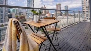 Un balcón con una mesa y sillas de madera, una planta en maceta y vistas a un paisaje urbano con edificios altos.