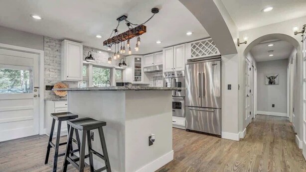 A modern kitchen with white cabinetry, a central island, stainless steel appliances, and a wooden floor. - Red Rock Farmhouse! Hot Tub, Fire Pit & Trails (Colorado Springs)
