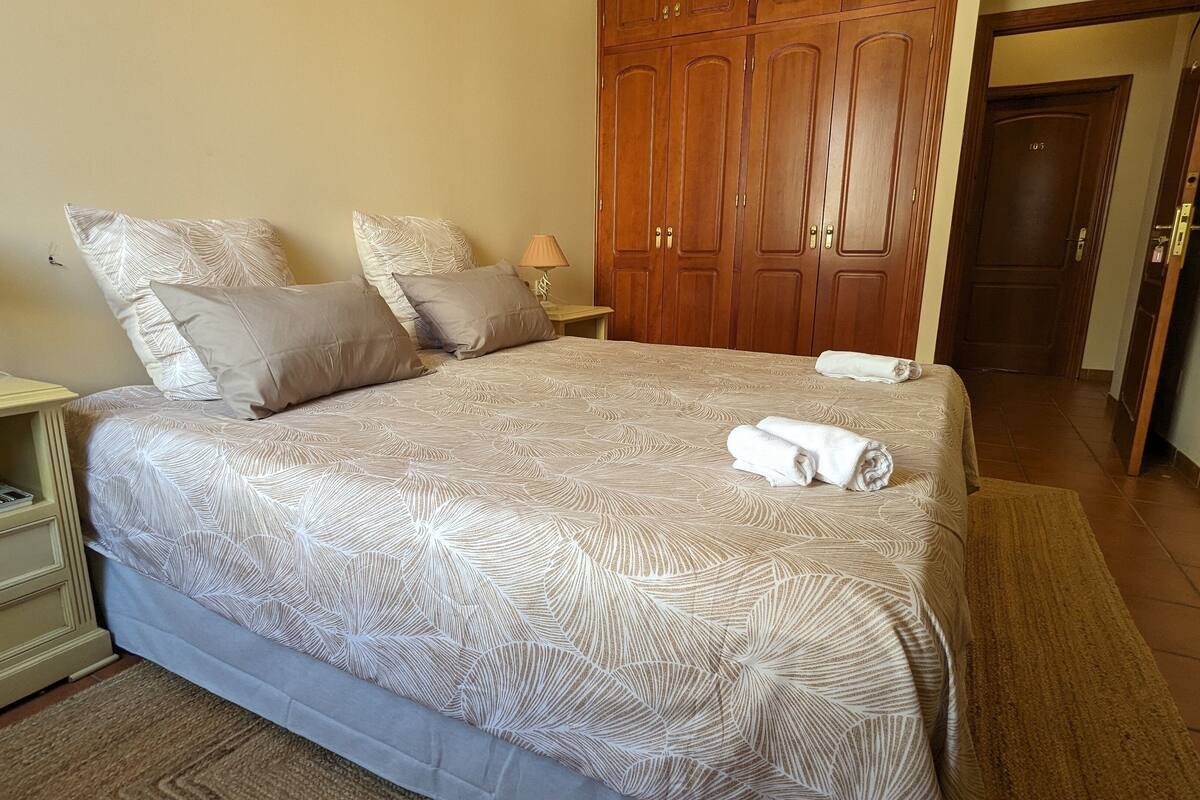 A neatly made bed with a patterned bedspread, two pillows, and two rolled towels on the bed.
