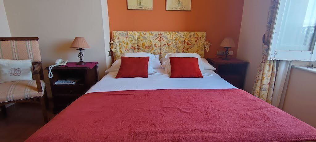 A hotel room with a bed, two red pillows, a nightstand with a lamp, a chair, and a window with curtains.