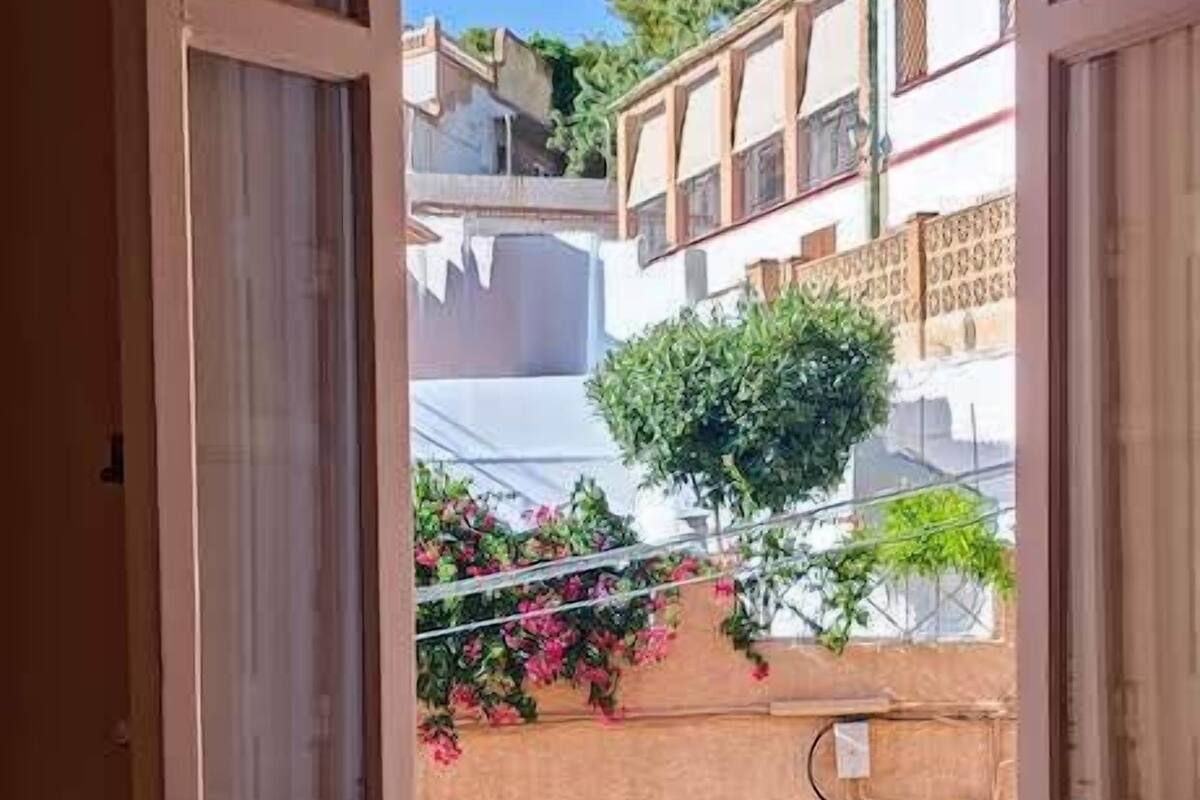 A bedroom with an open window overlooking a courtyard with buildings and greenery.