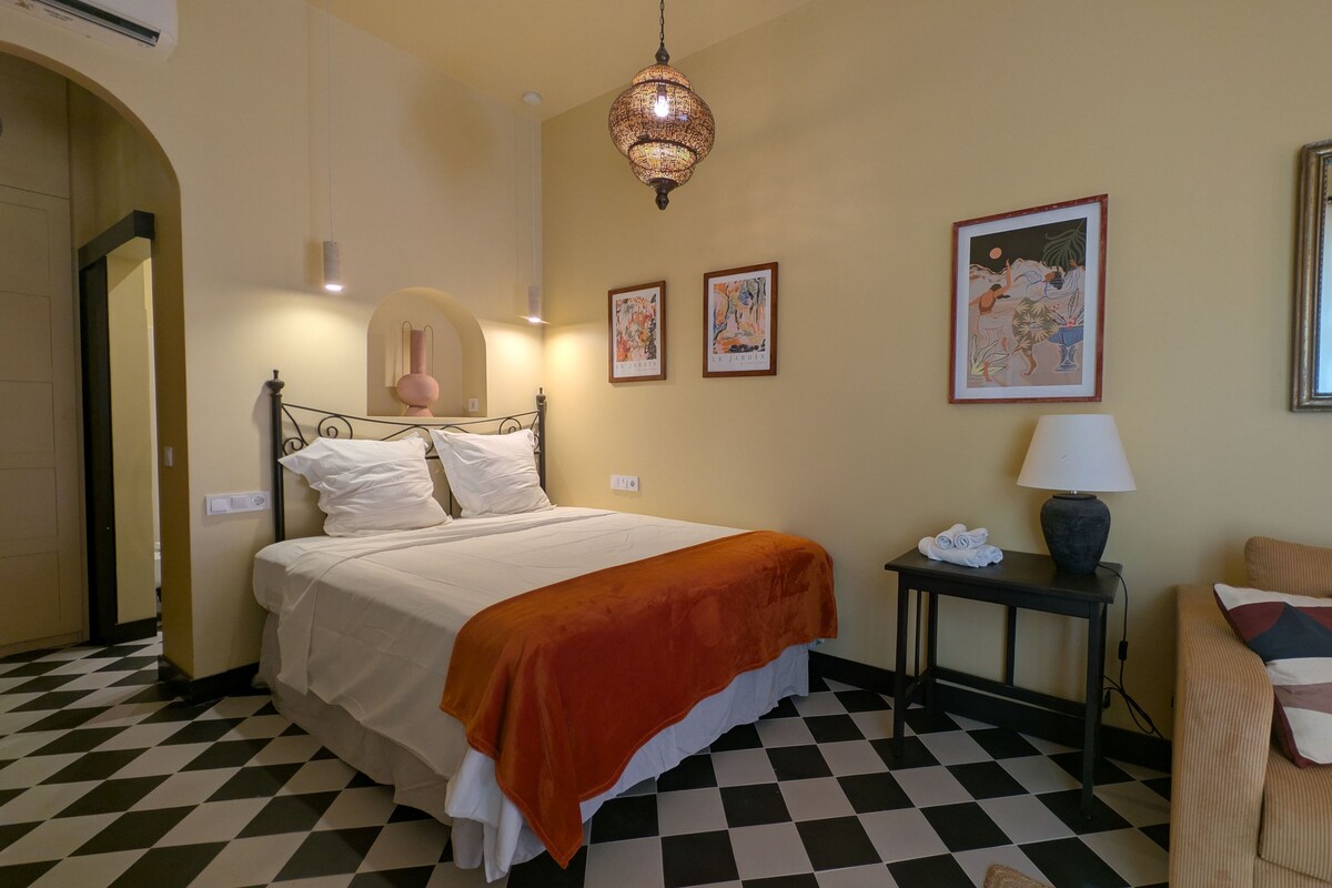 A hotel room with a checkered floor, a bed with white bedding and an orange throw, a bedside table with a lamp, and artwork on the wall.