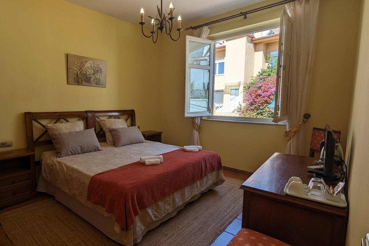 A bedroom with a bed, a bedside table, a desk with a computer, a window with a view of buildings and flowers, and a chandelier.